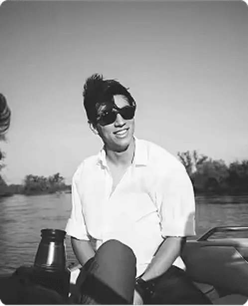 Man wearing sunglasses and a white shirt sitting on a boat with a river and trees in the background.