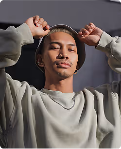Young man wearing a light-colored sweatshirt and bucket hat, holding the hat's brim with both hands against a dark background.