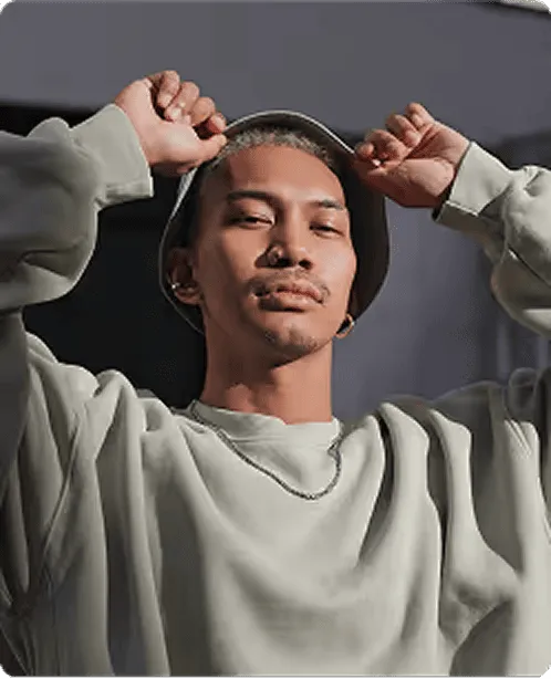 Young man wearing a light-colored sweatshirt and bucket hat, holding the hat's brim with both hands against a dark background.