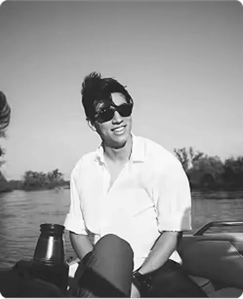 Man wearing sunglasses and a white shirt sitting on a boat with a river and trees in the background.