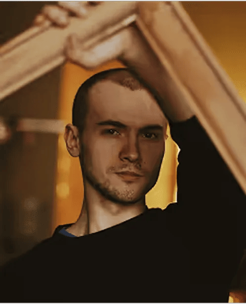 Young man with short hair and a serious expression holding a wooden frame in front of his face against a warm, blurred background.
