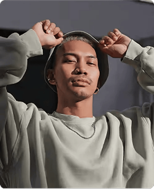 Young man wearing a light-colored sweatshirt and bucket hat, holding the hat's brim with both hands against a dark background.