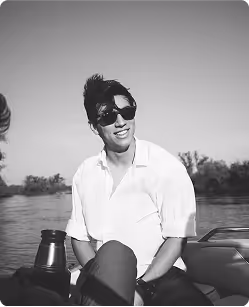 Smiling man wearing sunglasses and a white shirt sitting on a boat on a lake with trees in the background.