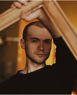 Portrait of a man with short hair and a trimmed beard looking directly at the camera, holding a wooden frame above his head.